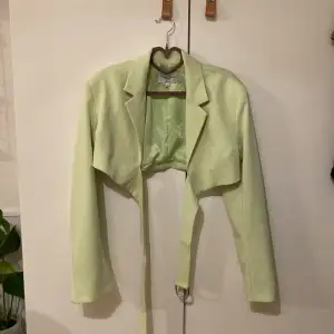 Croppad blazer i piggelin-grön färg från Angelica Blicks kollektion med nakd. Spänne som går från framsidan och fästs i ryggen. Använd 1 gång 