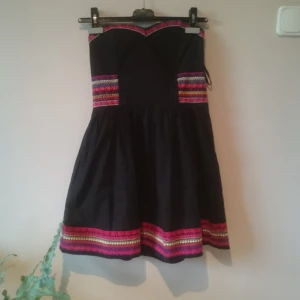  - Söt ärmlös klänning med färgglada broderier. Livet är en bustier och urringningen av modell sweetheart. Går att bära utan bh då den har en silikonkant och ståltrådsskenor, dock inga kupor. Den har smockresår i ryggen, dragkedja i sidan och två diskreta fickor.  Mycket fint skick, inga lösa trådar i broderiet etc.