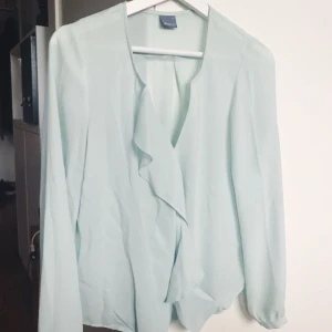  - Omlottblus med volang i ringningen. Svårfotad färg men den är en blek turkos färg.