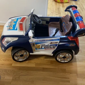 Policebil - Policebil till barn. Har laddare med sig. Vi har bytt batteri. Bilen har musik och man kan sätta på police ljus med knappen där bak. Har bälte och när man trycker med foten åker den. 