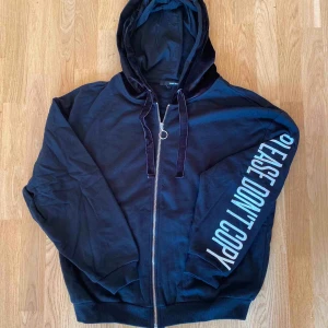  - Svart hoodie med textdetaljer på arm och rygg. Luva gjord i sammet. Helt oanvänd! 