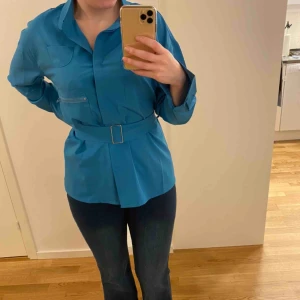 Skjorta med bälte - Luftig o tunt tyg med turkos färg. Blusen har fickor med dragkedja till på bröstet samt ett bälte i midjan med guldfärgat spänne. 