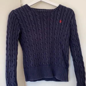 Kabelstickad tröja från Ralph lauren - Marinblå kabelstickad tröja från Ralph lauren med ett rött märke på. Köpt från barnavdelningen men skulle säga att det är en storlek xs men skulle även kunna passa xxs. Väl använd 🤍