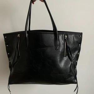 Väska  - Svart väska i medium storlek med nitar på sidorna. Har småfack på insidan och man kan stänga öppningen med en dragkedja. Använd vid ett tillfälle så är helt i nyskick. 