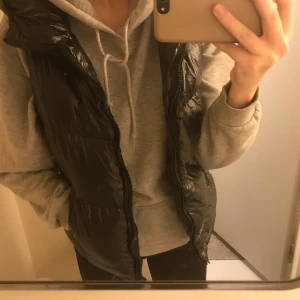 svart täckväst, ginatricot - Svart glansig täckväst med luva från ginatricot. Hyfsat tjockt material, ärvt av en kompis men aldrig använd av mig därav i väldigt bra skick. Köparen står för frakten. OBS: högsta bud 155kr+frakt