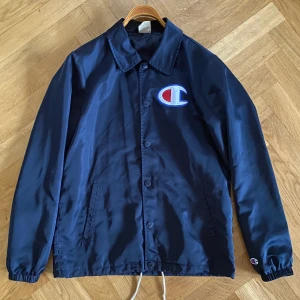 Champion coach jacka - Schysst coachjacka i polyester med en glansig marinblå finish. Lätt använd utan några fel.