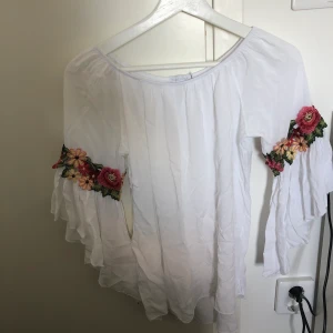 En off shoulder topp från Boohoo - En vit off shoulder topp från Boohoo i väldigt skönt somrigt material. Den består av fina blommiga detaljer längs armbågarna. Säljes på grund av flytt. 