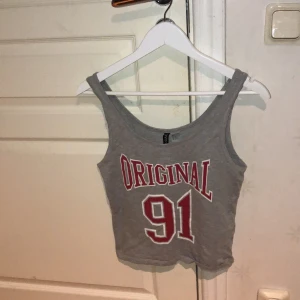 Ett grått linne med röd text -  Detta linne är grått med en röd basket text. Modellen är ganska kort och linnet är ganska tajt. Detta linnet passar jättebra till ett par grå eller svarta shorts. 