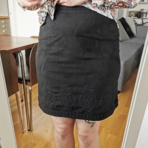 Kjol i mockaimitation  - Kjol i mockaimitation med broderier. Strl S. Midjemått 70 cm, längd 51 cm. Dragkedja i sidan. De stora blombroderierna är enbart på framsidan av kjolen, men i över- och nederkant fortsätter broderierna runt hela kjolen. Sparsamt använd, fint skick!
