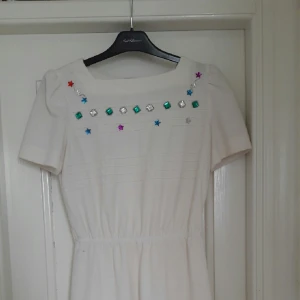  - Bli ägare till denna helt unika klänning! Vacker vintageklänning i krämig vit polyester. Dragkedja i ryggen och resår i midjan ger en jättefin passform! Spännande detaljer i form av knappar på ena axeln och färgglada glitterapplikationer över bysten. Mindre fläck nere vid midjan (se bild), i övrigt utmärkt skick. 