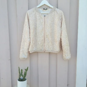  - Cremefärgad paljettjacka i bombermodell, fodring i satin. Gott begagnat skick. Inhandlad på second hand, men bara använd 1 gång.
