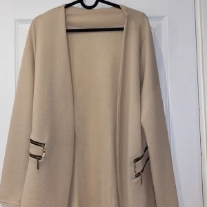 Beige kavaj - En längre beige kavaj i mjukt matterial med dragkedjor på sidan. Aldrig använd. Frakt tillkommer 