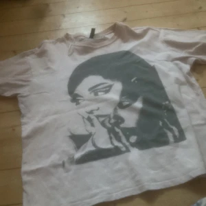 Vit t-shirt med tryck från Ariana Grande - Säljer en vit t-shirt med ett stort tryck av en person på framsidan. T-shirten är kortärmad och har en rund halsringning. Perfekt för fans av Ariana Grande!