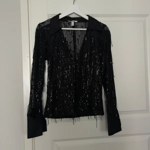 Svart glittrig blus - Snygg svart blus med glittriga detaljer och djup v-ringning. Blusen har långa ärmar och en klassisk krage, perfekt för nyår!!! 