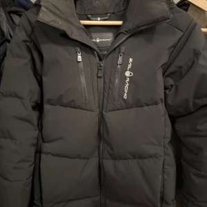 Svart dunjacka från Sail Racing - Säljer en svart dunjacka från Sail Racing, perfekt för kalla dagar. Jackan har en hög krage och dragkedja framtill, samt två praktiska bröstfickor med dragkedjor. Den är designad i Sverige och tillverkad i Vietnam, med en cool Sail Antarctica Expedition-märke på ärmen.