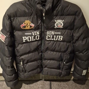 Svart dunjacka av Vinson Polo Club King jacket - Säljer en svart dunjacka från Vinson Polo Club med broderade detaljer och flaggor. Jackan har en dragkedja framtill och en huva med text. Perfekt för kyliga dagar! Varm jacka med stort tryck och snygga detaljer! Perfekt för höst/vinter. 100% polyesrer. Pris kan diskuteras.
