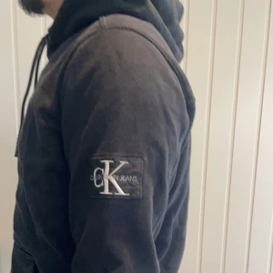 Svart hoodie från Calvin Klein - Snygg svart hoodie från Calvin Klein med en klassisk känguruficka och justerbar huva. Märkeslogga på ärmen ger en stilren touch. Perfekt för en avslappnad look.