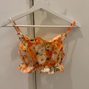 Blommig axelbands topp - Söt blommig topp i orange och vit med gröna detaljer. Axelbands design med volanger och elastisk midja för en bekväm passform. Perfekt för sommarens varma dagar.