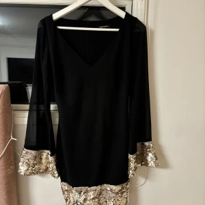 Svart klänning med paljettdetaljer från Explosion - Elegant svart klänning från Explosion med glittrande guldpaljetter vid ärmslut och nederkant. Klänningen har en v-ringad design och långa ärmar, perfekt för en festlig kväll.