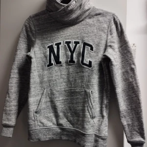 Grå hoodie med NYC-tryck - Säljer en grå hoodie med stort NYC-tryck på framsidan. Tröjan har en hög krage och en praktisk magficka. Perfekt för en avslappnad stil. Barn/ungdom storlek 146/152
