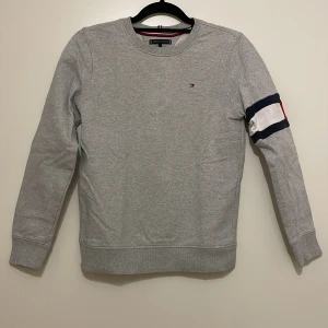 Grå sweatshirt från Tommy Hilfiger - Säljer en stilren grå sweatshirt från Tommy Hilfiger med ett diskret logotypbroderi på bröstet. Tröjan har långa ärmar med en röd och blå randdetalj på ena ärmen. Finns dock en röd fläck på höger ärm. Perfekt för en avslappnad look.