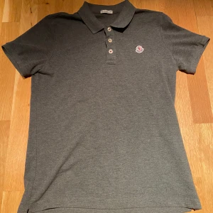 Grå pikétröja från Moncler - Snygg grå pikétröja från Moncler med klassisk krage och knappslå. Tröjan har en broderad Moncler-logga på bröstet. Perfekt för en stilren och avslappnad look. Storlek M men passar både M och S