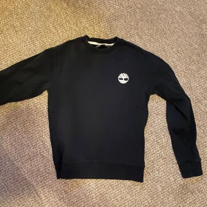 Svart sweatshirt från Timberland - Säljer en stilren svart sweatshirt från Timberland med deras ikoniska logga på bröstet. Tröjan har långa ärmar och en klassisk rund halsringning. Perfekt för en avslappnad stil.