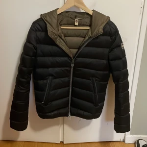 Svart pufferjacka från Colmar - Snygg svart pufferjacka från Colmar med huva och dragkedja. Perfekt för kyliga dagar med sin quiltade design och långa ärmar. Jackan har en stilren look och är idealisk för både stad och natur. Skriv om frågor och kan gå ner vid snabb affär