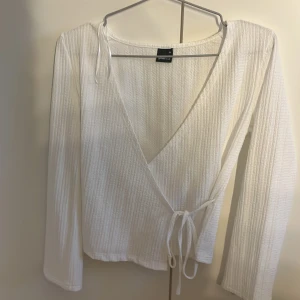 Vit omlottopp från Gina - Säljer en stilren vit omlottopp från gina Toppen har långa ärmar och en knytning i midjan för en justerbar passform. Materialet är ribbat vilket ger en fin textur och extra stilpoäng. Perfekt för både vardag och lite finare tillfällen.