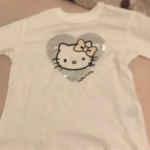 Vit Hello Kitty t-shirt från H&M - Söt vit t-shirt från H&M med ett stort Hello Kitty-tryck i silver och rosa. Perfekt för alla Hello Kitty-fans! T-shirten har korta ärmar och är gjord i mjukt bomullsmaterial.