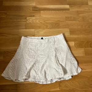 Vit spetskjol från H&M - Säljer en söt vit spetskjol från H&M i storlek M. Kjolen har ett vackert spetsmönster och en knappdetalj i midjan. Perfekt för varmare dagar och ger en romantisk touch till din outfit.