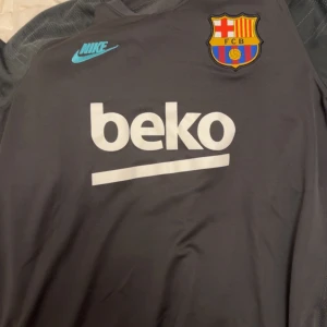 Grå Nike Dri-FIT FCB tröja - Säljer en grå Nike Dri-FIT tröja med FCB-logga och Beko-tryck. Perfekt för fotbollsträning med sin andningsbara design. Tröjan har korta ärmar och en sportig look.