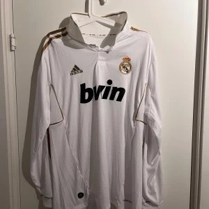 Vit långärmad Real Madrid tröja från Adidas - Säljer en vit långärmad Real Madrid fotbollströja från Adidas med klubbens emblem och sponsorlogga på framsidan. Tröjan har gulddetaljer på axlarna och ärmarna. Perfekt för fans av laget!