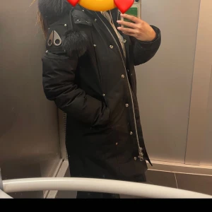 Svart parkajacka med pälsdetaljer - Snygg svart parkajacka från Moose Knuckles med pälsdetaljer på huvan. Jackan har en lång passform och är utrustad med både dragkedja och knappar. Perfekt för kalla dagar.pris kan diskuteras.