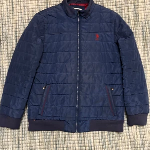 Blå puffer jacka från U.S. Polo Assn. - Säljer en snygg blå puffer jacka från U.S. Polo Assn. med röd broderad logga på bröstet. Jackan har dragkedja och ribbade muddar vid ärmslut och nederkant. Perfekt för kyligare dagar!