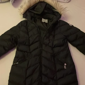 Svart dam moncler jacka - Snygg svart dunjacka med huva och pälsdetaljer. Jackan har dragkedja och praktiska fickor med dragkedjor. Perfekt för kalla dagar. Den är en fin present att ge till nån nära.