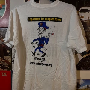  vintage t-shirt med tryck från Unió de Joves - Vit t-shirt med tryck både fram och bak. Framsidan har texten 'Fumem-ho Legal!' i svart. Baksidan har en tecknad figur i blå uniform med texten 'Legalitzem les drogues toves' och 'www.uniodejoves.org'.