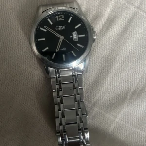 Silverfärgad klocka från GHW - Stilren klocka från GHW med svart urtavla och datumvisning. Boetten och armbandet är i silverfärgat metall. Perfekt för en elegant look.