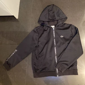 Svart hoodie från Lacoste - Säljer en stilren svart hoodie från Lacoste med dragkedja och logotyp på bröstet. Hoodien har långa ärmar och ribbade muddar. Perfekt för en avslappnad stil. Köptes för 800kr, men säljes för 400kr. Helt ny aldrig använts.
