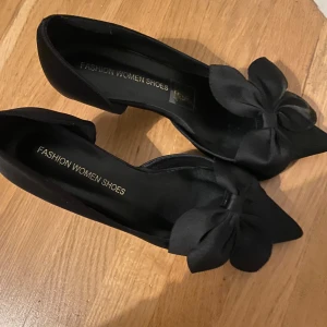 Svarta klackar med rosett - Eleganta svarta ballerinaskor med en stor dekorativ rosett på tån. Perfekta för en stilren look.