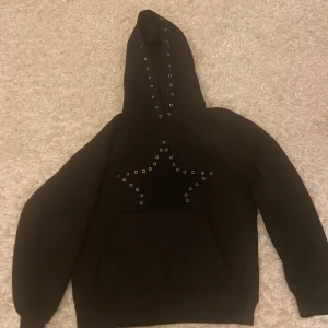 Svart hoodie med stjärndetalj - Snygg svart hoodie med en stor stjärna av nitar på framsidan. Huvan har också nitar för en extra cool look. Perfekt för en avslappnad stil.