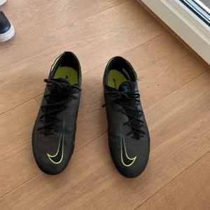 Svarta Nike fotbollsskor - Snygga svarta Nike fotbollsskor med gula och blå detaljer. Skorna har snörning och en modern design med Nike-loggan på sidan. Storlek 46 passar även 45