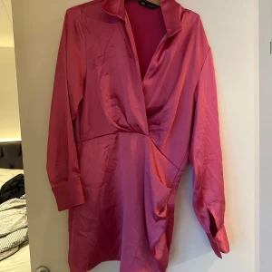 Rosa klänning från Zara - Säljer en snygg rosa klänning från Zara i satin med lång ärm och en elegant omlott-design framtill. Perfekt för en chic look! Finns lite trasiga sömmar vilket syns på bilderna