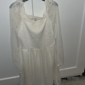 Vit klänning med spetsdetaljer - Charmig vit klänning med spetsdetaljer och långärmad design. Den har en luftig passform och ett romantiskt utseende med volanger vid axlarna. Perfekt för en somrig look.