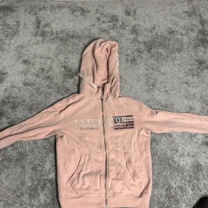 Rosa hoodie från Lexington - Mysig rosa hoodie från Lexington med dragkedja och tryck på bröstet. Perfekt för en avslappnad stil. Den har en praktisk huva och fickor framtill.