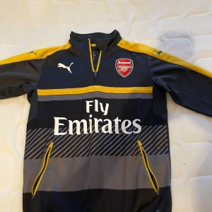 Arsenal tröja från Puma - Snygg Arsenal tröja från Puma i mörkgrått med gula detaljer och klubbens emblem på bröstet. Tröjan har en halv dragkedja och långa ärmar, perfekt för fotbollsfans. Fly Emirates-logga pryder framsidan. Passar typ S