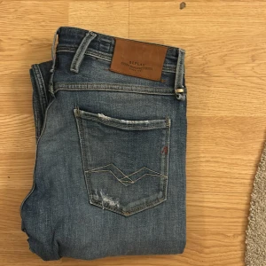 Blå jeans från Replay - Säljer ett par snygga blå Replay Anbass jeans i slim fit. De har en cool slitning och en klassisk femficksdesign. Perfekta för en avslappnad stil.