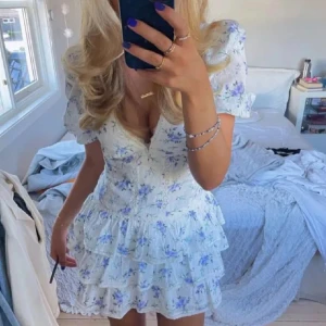 Blommig vit och blå klänning - Charmig kortklänning med blommigt mönster i vitt och blått. Klänningen har volanger och en v-ringad design med korta ärmar. Perfekt för sommarens festligheter. Aldrig använd själv men köpt på Plick 