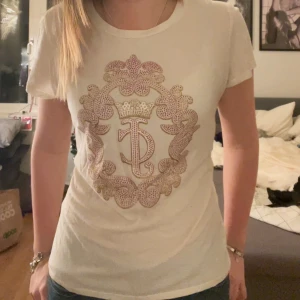 Vit t-shirt från Juicy Couture - Snygg vit t-shirt från Juicy Couture med ett stort dekorativt emblem i mitten. Perfekt för en avslappnad stil med en touch av glamour.