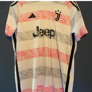 Juventus fotbollströja från Adidas - Snygg Juventus fotbollströja från Adidas med korta ärmar. Tröjan har ett randigt mönster i vitt, svart och rött med klubbens logga och sponsortryck på framsidan. Perfekt för fans som vill visa sitt stöd!